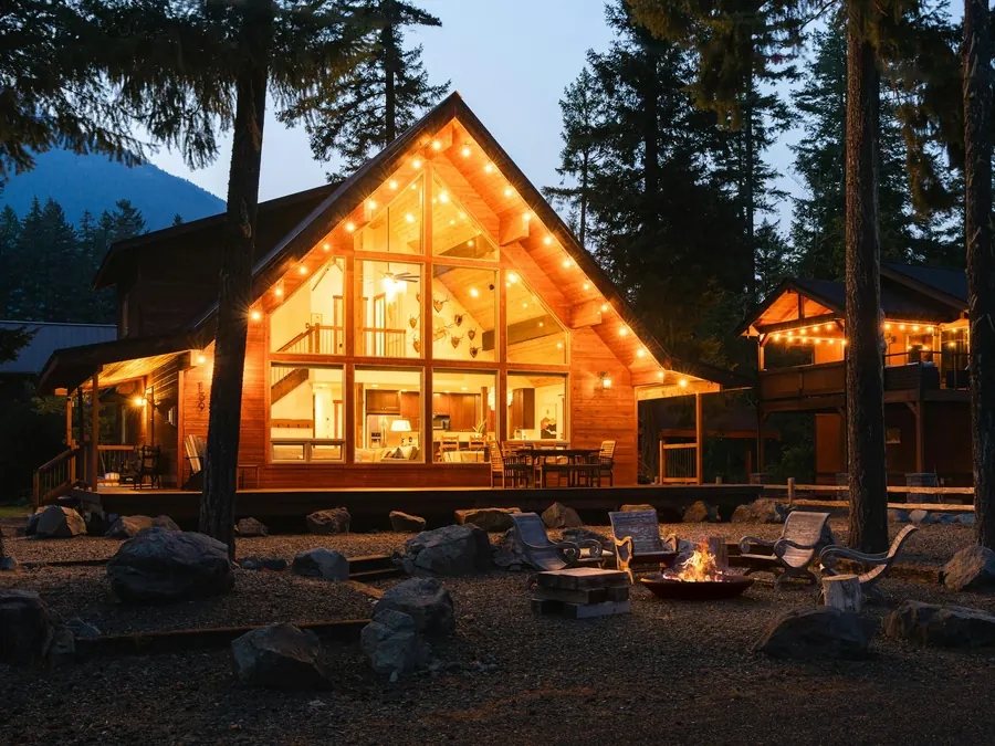 A-frame mountain cabin with glowing floor-to-ceiling windows and a fire pit among pines at dusk